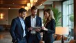 © Viktor - Three business professionals in suits discuss strategy with a tablet in a modern hotel lobby. Diverse team collaborates, planning growth in a luxury indoor space.