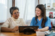 © VStudio - Two women interact at a wooden table. An older woman with short gray hair and glasses uses a tablet. Professional nurse in blue scrubs consults her. They discuss home care and retirement planning