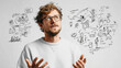 © Anchanar S - Thoughtful man in glasses and white sweater gestures while discussing creative ideas, with hand drawn sketches on whiteboard behind him, illustrating brainstorming session
