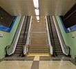 © teracreonte - Escalera central rodeada por dos escalera mecánicas (de subida y de bajada), en un pasillo con suelo con pavimento con guía táctil, en una estación de metro