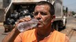 © Olga Ко - Exhausted sanitation worker drinking water from a plastic bottle during an extreme summer heatwave, standing in front of a garbage truck
