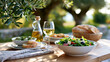 © YURII - Fresh Mediterranean salad served on a wooden table outdoors with olive oil, white wine, and bread in a sunny garden setting surrounded by greenery