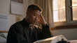 © forenna - Young seminarian sits alone in dormitory room at night, open theology textbook in lap, hand pressing temple in exhausted uncertainty, wall calendar and acceptance letter from