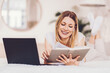 © Prostock-studio - Smiling Caucasian Woman Enjoying Reading Book While Working On Laptop At Home, Sitting Comfortably On Bed In Cozy Indoor Setting, Representing Relaxed Lifestyle And Cozy Atmosphere