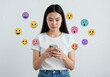 © Jaetin - Young Asian woman looking down at her mobile phone surrounded by colorful floating holographic emoji faces showing mixed positive and negative human emotions on a plain white studio background.