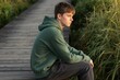 © Lubov - Young man sits on a wooden boardwalk in green hoodie. Boy looks thoughtfully at grassy nature. Teenager reflects in quiet solitude on path. Peaceful, outdoor mood.