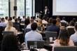 © Kevin - Man in a suit giving a presentation to a large group of people in a conference room