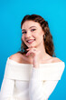 © deagreez - Portrait of a smiling young woman wearing a white off-shoulder sweater and hoop earrings posing confidently against a bright blue background