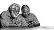 © Christelle - Black and white portrait of a senior Aboriginal couple looking at a smartphone screen with curious expressions while sitting at a wooden table against a white background.