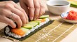 © Konika - Person hands preparing fresh sushi roll with avocado and salmon on a bamboo mat in kitchen
