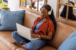 © wavebreak3 - African American adult woman sitting cross-legged on sofa at home using laptop wearing headphones