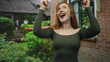 © Krakenimages.com - Redhead woman pointing both index fingers upward with bare shoulders visible, smiling in front of a brick building and lush garden; joy celebration.