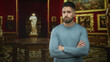 © Krakenimages.com - Man with beard arms crossed beside classical statue and gilded paintings inside ornate museum building; art contemplation skepticism.
