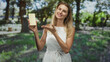 © Krakenimages.com - Woman holding white chocolate bar in one hand and gesturing open palm with other, wearing white dress in forest; joyful delight.