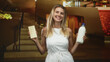 © Krakenimages.com - Woman with blonde hair holds a white chocolate bar and a milk bottle, smiling and presenting both items in a building lobby staircase; joy indulgence choice.