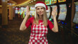 © Krakenimages.com - Woman in santa hat and red checkered apron raises hands palms up near face inside casino slot machines building; festive.