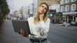 © Krakenimages.com - Woman holding laptop with chin on fist and hand covering mouth on urban street; tired remote work focus.