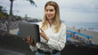 © Krakenimages.com - Blonde woman holding laptop with hand to mouth gesture at the beach, wearing white shirt and jeans, standing near shoreline umbrellas and rocks; surprise remote work.