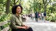 © Xenia - Smiling Black woman sitting on a park bench looking away. African American female relaxing outdoors on a sunny summer day. Urban leisure and wellbeing concept