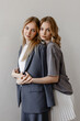 © Kaplitskaya Love - Two young women pose closely together against a plain light background, one wearing a tailored dark suit and the other in a gray top with a white pleated skirt