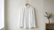 © gianfelixr - A crisp white button-down shirt hangs on a wooden hanger against a neutral wall, with a small plant on a wooden shelf to the right.