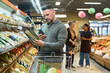 © pressmaster - Man comparing grocery product details on smartphone while shopping in supermarket. For retail service marketing