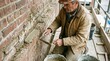 © Nuttawut - Bricklayer Restoring Wall: A skilled bricklayer, focused and weathered, meticulously repairs an old brick wall with cement, his hands calloused and experienced.