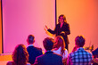 © Lustre Art Group  - Business speaker presenting to audience in neon conference room. Concept of public speaking, corporate training, seminar education, leadership communication and presentation skills.