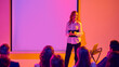 © Lustre Art Group  - Female presenter speaking to seated audience near blank projection screen. Concept of corporate seminar, business education, leadership development, workshop training and communication skills.