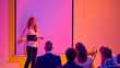 © Lustre Art Group  - Female presenter speaking to audience beside blank screen in neon seminar room. Concept of business seminar, workshop training, public speaking, leadership and corporate education.