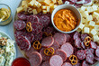 © Oksana - A round sharing board with salami slices cheese cubes pretzels walnuts and a red pepper dip on a white table a festive charcuterie platter for entertaining.