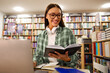 © Home-stock - Positive female student reading book in front of laptop in university library sitting at desk, preparing for project and searching information