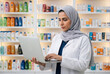 © Meow Creations - Female muslim pharmacist in a hijab and white lab coat using a laptop in a brightly lit pharmacy