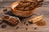 Cinnamon sticks in wooden bowl with ground cinnamon powder and various spices scattered on a rustic surface
