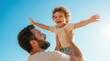 © Masque - A happy bearded father lifting his curly haired toddler son high against a clear blue sky.
