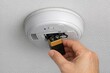 © Ева Поликарпова - A person is shown holding a battery in front of a smoke detector, possibly for replacement or maintenance