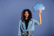 © standret - Young African woman with curly hair holds a blue speech bubble cutout against a solid purple background, wearing a denim shirt and casual attire