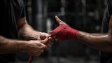 Two pairs of hands are shown in close-up as one person helps another prepare for combat sports by applying a protective red wrap in a high-contrast lighting setting