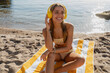 © mary_markevich - attractive blond woman in bikini sunbathing on beach in Italy listening to music in headphones smiling happy dressed in yellow swimwear, sitting on towel on sand, summer season fashion trend style