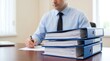 © CuteBee - Professional businessman sitting at desk signing documents with stack of blue ring binders in foreground showing heavy workload and corporate administration tasks in bright office