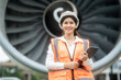 © Thitaporn - Smiling female engineer in safety vest and helmet holding a tablet in front of a large jet engine at an airport, showing aviation work, inspection, maintenance and travel industry.