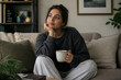 © mila103 - Young woman holding mug sitting on sofa in living room