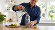 © LM - Senior man pouring water from a jug into a reusable glass bottle in a bright modern kitchen with fresh mint and lemon on the counter