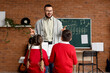 © Pixel-Shot - Music teacher guiding young students in piano lesson with musical staff on chalkboard in classroom, the instructor stands in front of two children, a boy and a girl