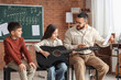 © Pixel-Shot - Young children learning guitar from male teacher in music class with chalkboard notes, the classroom setting includes a chalkboard with musical notes, emphasizing the educational environment