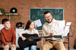 © Pixel-Shot - Music teacher guiding young girl playing acoustic guitar with boy observing in classroom setting, the teacher holds sheet music, demonstrating notes to the focused student