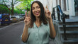 © Krakenimages.com - Hispanic woman brunette smiling and showing peace sign with both hands on a city street stoop near parked cars; playful confidence.