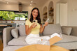 © Home-stock - Happy woman looking at smartphone with smile and holding skincare serum while opening delivery box filled with cosmetic products on sofa