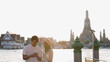 © blueringmedia - Young Asian Couple Using Smartphone by Riverside Temple at Sunset