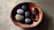 © Honey - A wooden bowl containing smooth dark massage stones and a small glass bottle of essential oil for relaxing spa treatments on a textured brown fabric background